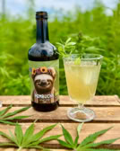 Eine Flasche mit einem Faultier-Etikett und ein Glas mit einem Getränk stehen auf einem Holztisch, umgeben von grünen Blättern und Pflanzen im Hintergrund.