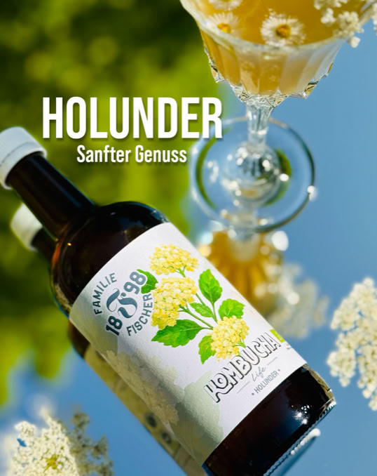 Eine Flasche mit einem Etikett, das Holunderblüten zeigt, liegt neben einem gefüllten Glas auf einem spiegelnden Untergrund. Im Hintergrund ist unscharf grünes Laub zu sehen. Text: "Holunder Sanfter Genuss".