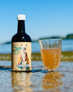Eine Flasche mit Etikett und ein Glas mit hellbrauner Flüssigkeit stehen auf nassem Sand vor einem unscharfen Hintergrund mit Wasser und blauem Himmel.