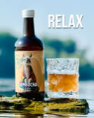 Eine Flasche Kombucha und ein Glas mit Getränk stehen auf einem Felsen, im Hintergrund unscharfe Natur. Oben rechts steht der Text "RELAX".