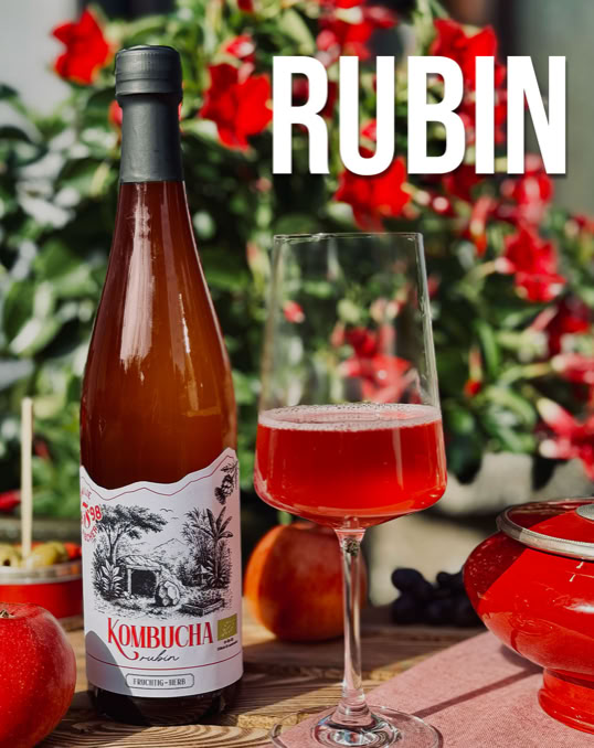 Eine Flasche und ein Glas mit rotem Getränk auf einem Tisch, umgeben von roten Blumen und Äpfeln, mit dem Wort "RUBIN" im Hintergrund.