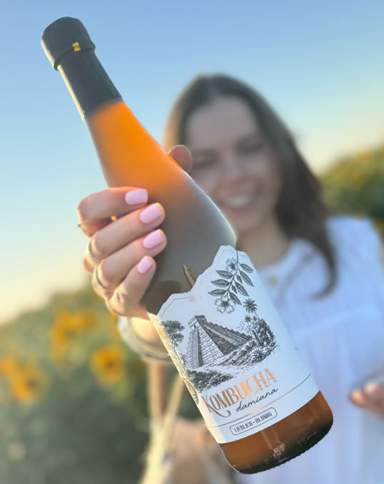 Eine Person hält eine Weinflasche in der Hand, im Hintergrund sind Sonnenblumen und ein blauer Himmel zu sehen.