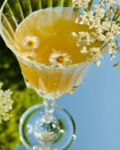 Kristallglas mit gelbem Getränk, dekoriert mit kleinen weißen Blüten und schwimmenden Blüten im Getränk.