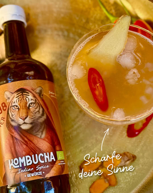 <!-– Eine Flasche Kombucha mit einem Tiger auf dem Etikett neben einem Glas mit Getränk, Eiswürfeln, einer Chilischote und einem Ingwerstück. Text: "Scharfe deine Sinne". –>