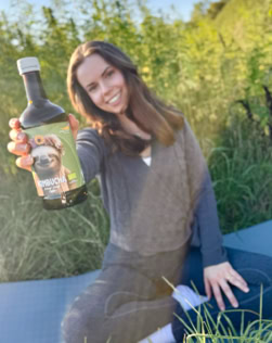 Eine Frau sitzt im Gras und hält eine Flasche mit einem Etikett, das einen Hund zeigt.