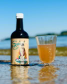 Eine Flasche mit Etikett und ein Glas mit hellbrauner Flüssigkeit stehen auf nassem Sand vor einem unscharfen Hintergrund mit Wasser und blauem Himmel.