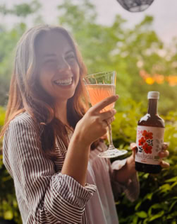 Eine lächelnde Person hält ein Glas mit rosa Getränk in der einen Hand und eine Flasche mit Etikett in der anderen, im Hintergrund grüne Pflanzen.