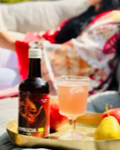 Eine Flasche Kombucha und ein Glas mit rosa Getränk auf einem Tablett neben einem Apfel und einer Birne, im Hintergrund eine Person auf einem Sofa.