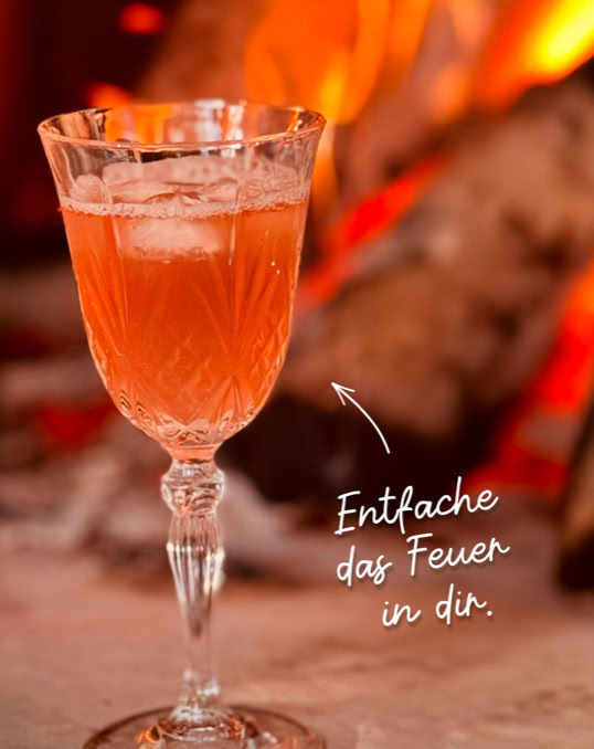 Kristallglas mit orangefarbenem Getränk und Eiswürfeln vor einem Kaminfeuer, daneben der Text "Entfache das Feuer in dir."