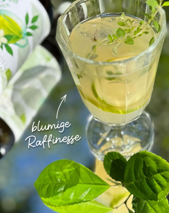 Ein Glas mit einem hellen Getränk und Kräutern, daneben grüne Blätter und eine Flasche mit einem Zitronenmotiv.