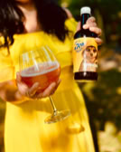 Eine Person in einem gelben Kleid hält ein Glas mit einem Getränk und eine Flasche mit einem Etikett, das einen Hund zeigt.