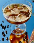 Ein Glas mit einem braunen Getränk und Eiswürfeln, umgeben von Kaffeebohnen auf einer reflektierenden Oberfläche.