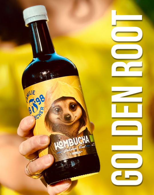 Eine Hand hält eine Flasche Kombucha mit einem Etikett, das ein Erdmännchen zeigt. Daneben steht der Text "GOLDEN ROOT".
