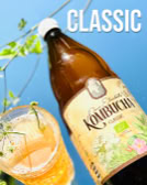 Braune Kombucha-Flasche und gefülltes Glas auf blauem Hintergrund mit grünem Zweig, Text "CLASSIC" oben rechts.